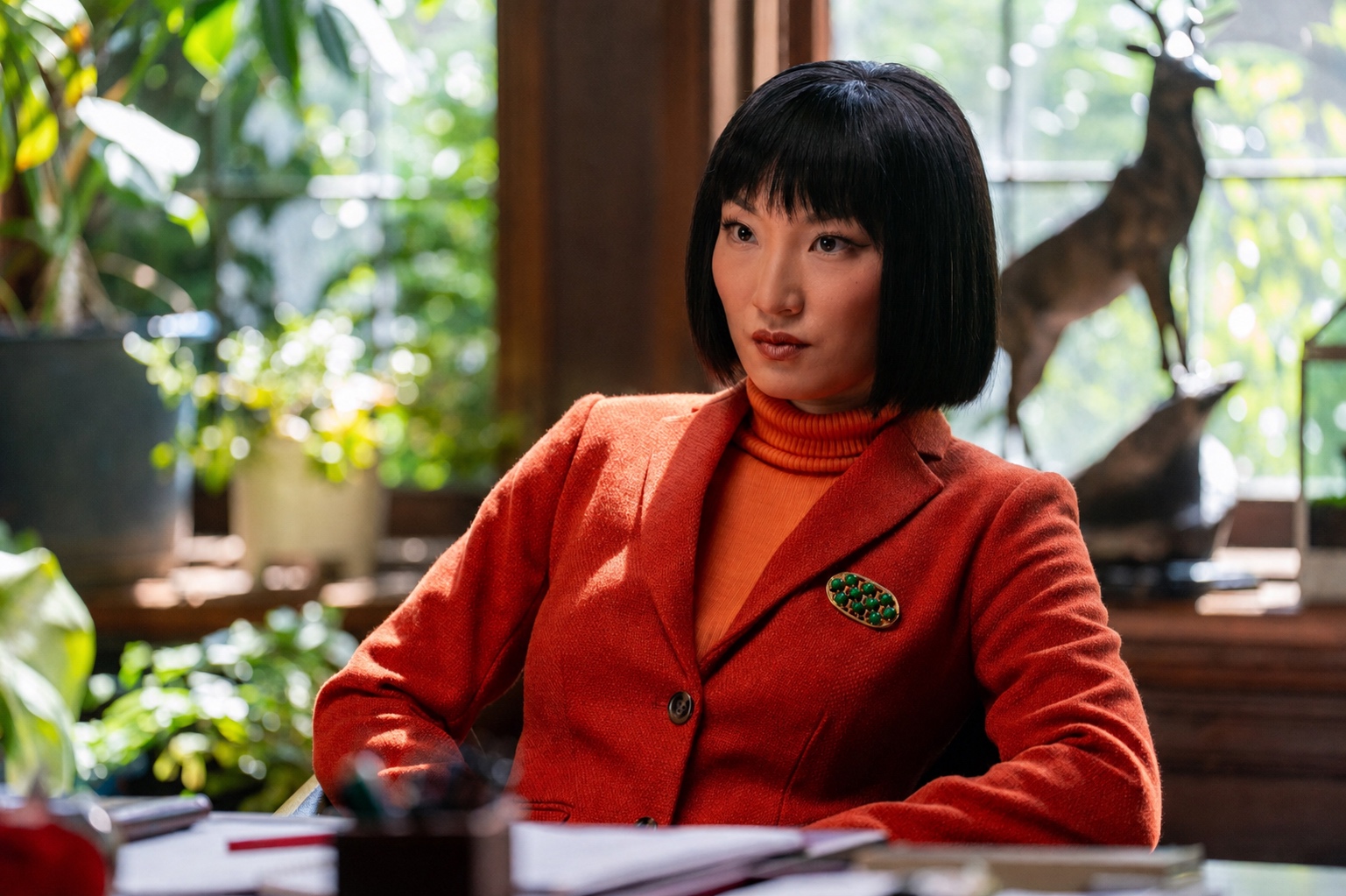 A woman in a red wool suit and orange turtleneck seated at a desk, His & Hers