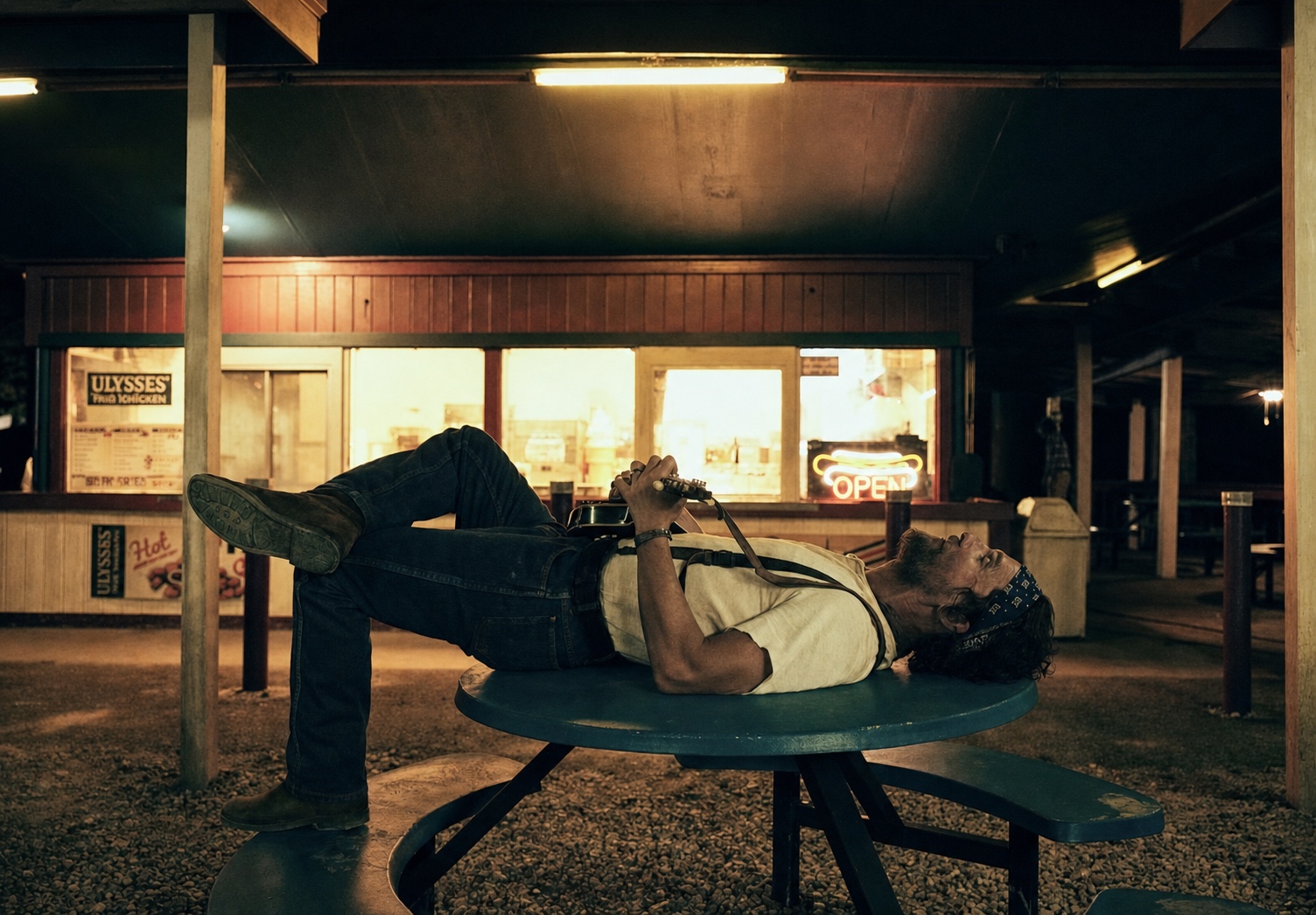 Matthew McConaughey lying on a picnic table with a mandolin outside a lit roadside diner at night, The Rivals of Amziah King